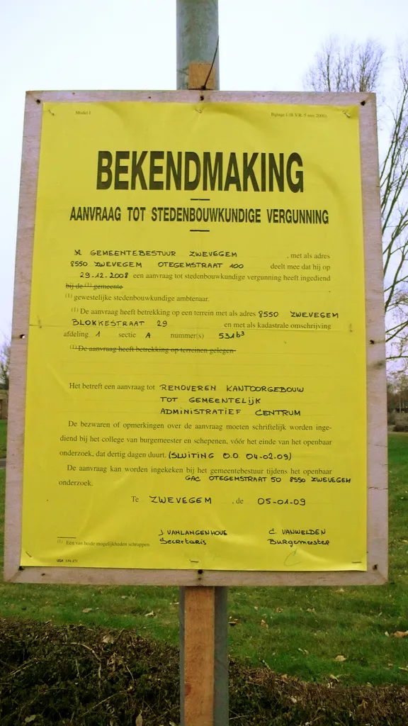 Een geel bekendmakingsbord op een houten paal in het gras: aanvraag tot stedenbouwkundige vergunning in Zwevegem