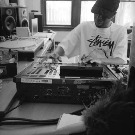 Zwart-witfoto van J Dilla met pet en Stussy-shirt, spelend op een sampler in een studio met draaitafels