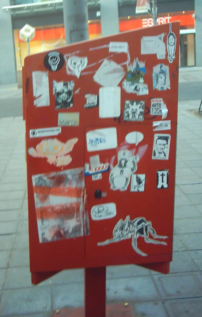Een rode brievenbus volgeplakt met stickers van straatartiesten, in een winkelstraat