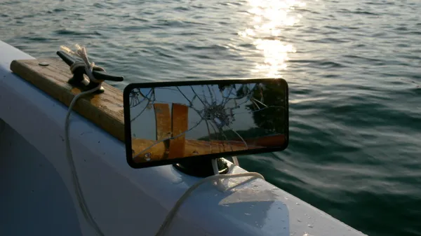 Een gebarsten achteruitkijkspiegel op de reling van een witte boot, met glinsterend water op de achtergrond